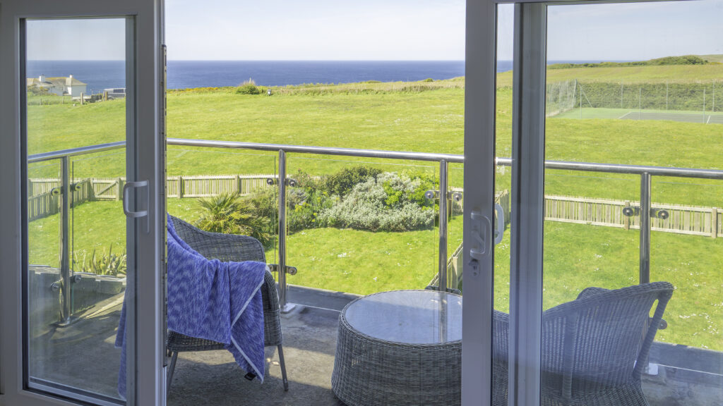 Villa 21. View of the balcony and surrounding areas from inside the villa. Balcony with two chairs and a coffee table. View below of villa garden, Tennis court and Tennis court lawns. Ocean view.