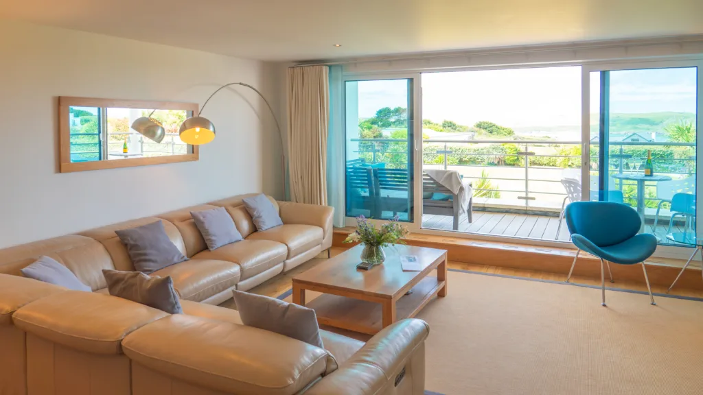 Apartment 105 living area and balcony view. The living area has an L shaped sofa and coffee table. The sliding glass doors are open leading out onto the balcony. The balcony has benches and a chair and table on it. Off the balcony is the view of the gardens below.