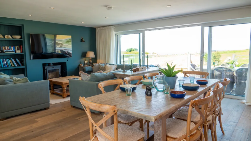 Villa 25 dining and living area. L shaped sofa and smaller sofa around the coffee table in the living area. Log burner below the TV on the wall. The dining table with chairs at the front of the image. Sliding glass doors are open leading to the balcony. View of the tennis court lawn in the background.