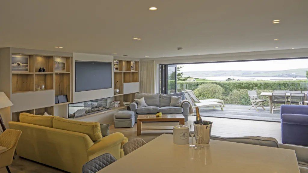 Villa 31 living area and balcony. View of the room from the kitchen island. A large living area with two sofas on the left of the room and an arm chair on the right. A pouffe and a coffee table in the middle of the seating area. Behind the living area the sliding glass doors are open and lead out onto the balcony. The balcony is furnished with two sunlounges and a dining table with chairs. The view of the sea and headland in the distance.