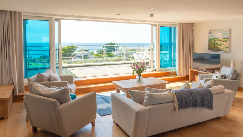 Apartment 308 living area. Three arms chairs and a sofa surrounding a wooden coffee table in the living area. The living area is looking out onto the balcony and view through the glass sliding doors that are open. The balcony has a dining table and chairs on. The view of the gardens and other buildings below. The ocean is in the distance.