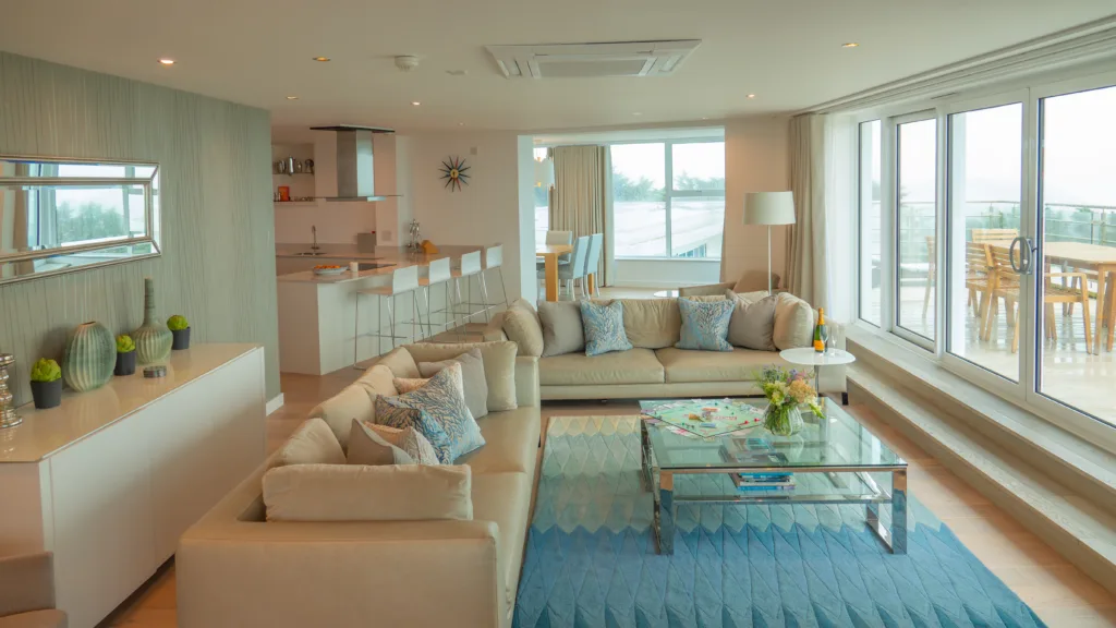 Apartment living area. Two sofas and an arm chair around the glass coffee table in the living area. B hind the sofas is the breakfast bar/island of the kitchen. There is also the entrance way to the dining area.