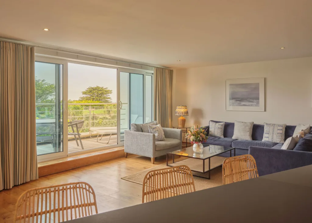 View of the living area from the kitchen. The living area is on the right side. of the room with an arm chair and L shaped sofa surrounding the glass coffee table. Behind the living area are balcony doors which are open and leading out onto the balcony. The balcony is furnished with 2 sun lounges and a small dining table and chairs. A view of the gardens below in the distance.