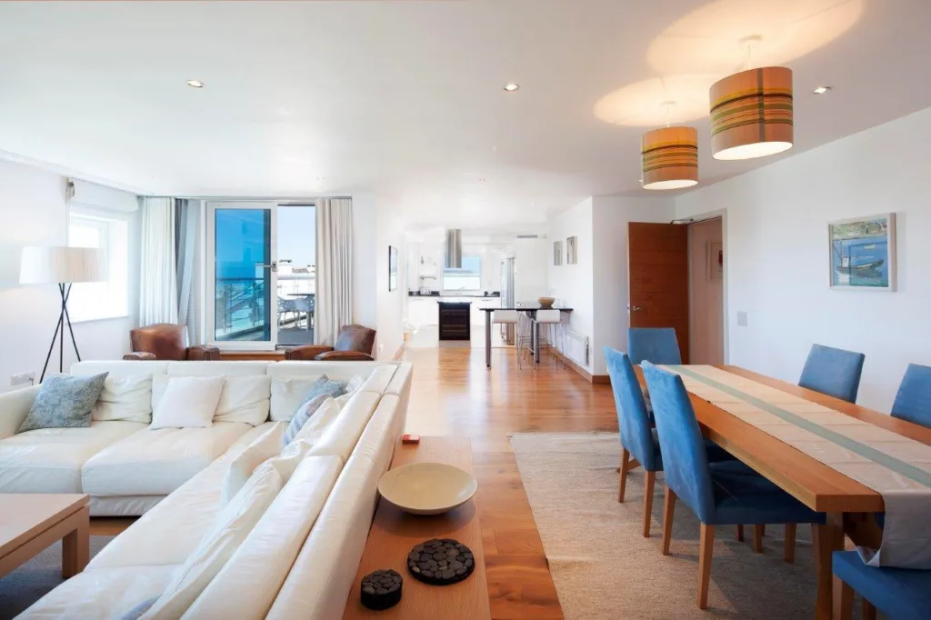 Apartment 201 living, kitchen an dining area. L shaped sofa and coffee table in the left corner of the image. Behind the L shaped sofa are two arm chairs near the door leading to the balcony. The kitchen is at the back of the image with a breakfast bar and two stools at the front of the kitchen area. On the right of the image is a door to the other areas of the apartment. There is a dining table and chairs in the front right corner of the image.