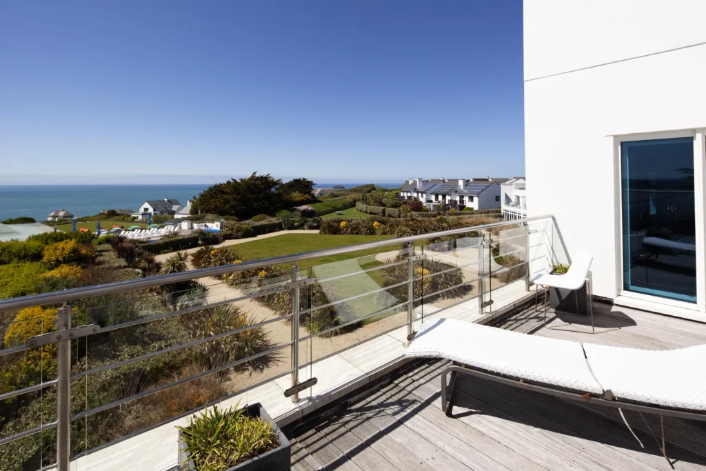 Apartment 210 balcony and view off the balcony. Balcony furnished with a sunlounge and a chair. There is a door leading back into the apartment. There is a view of the gardens and seaside area and surrounding areas below off the balcony. The ocean is in the distance.
