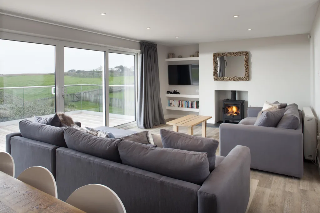 Villa living area. L shaped sofa and another sofa with a coffee table facing the TV in the corner on the wall. There is a fire place. The living area also looks out onto the balcony and the tennis court and tennis court lawn below that.