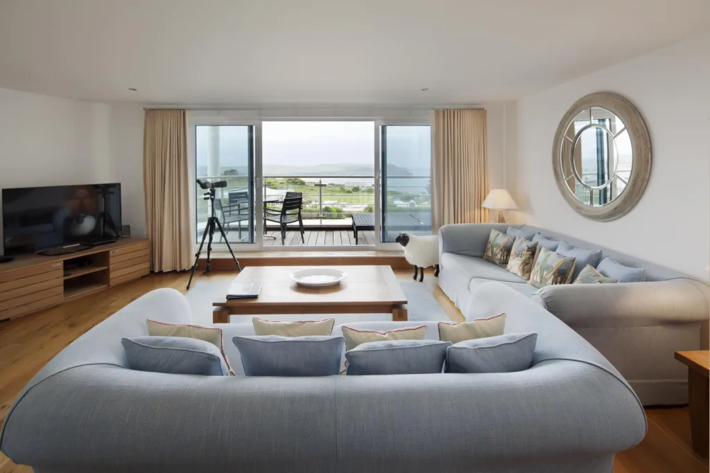 Apartment 206 living area. Two sofas with a wooden coffee table in front. TV on stand to the left of the image. The living area looks out onto the balcony and view beyond that. The balcony is furnished and has the view of the gardens and seaside area below and the sea and headland in the distance.